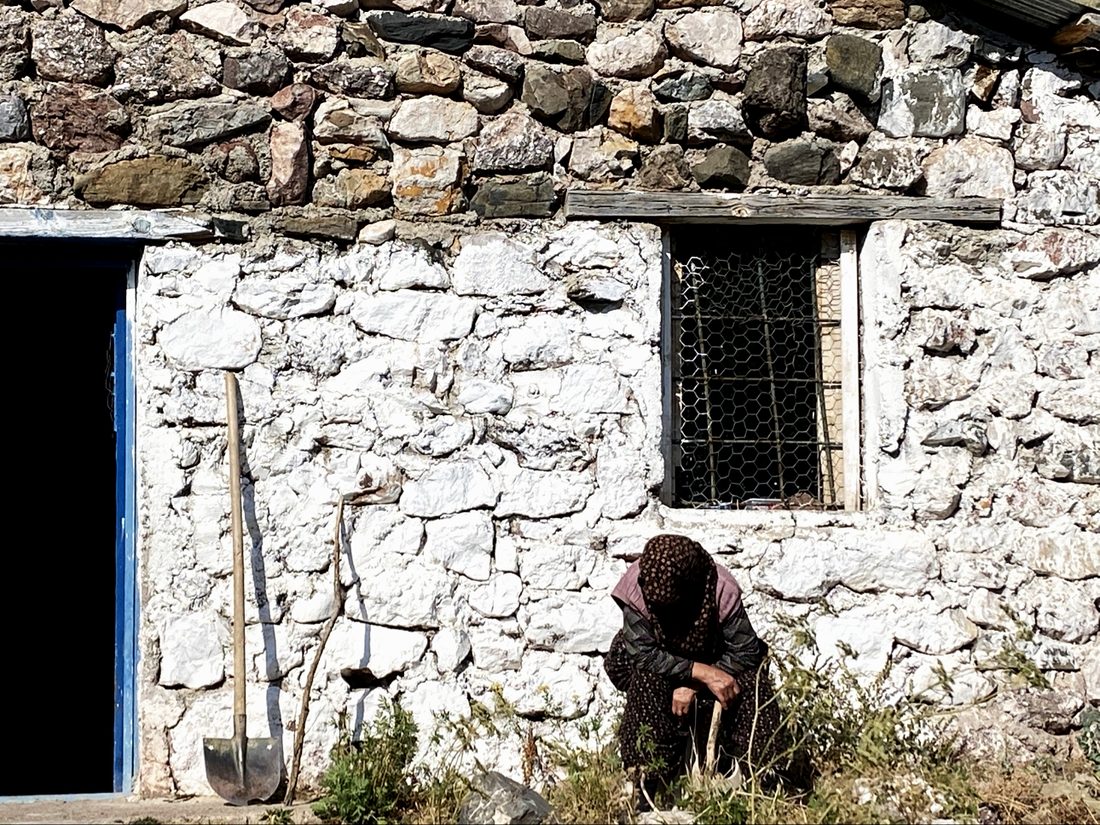 An old woman resting against the whitewashed stone wall of a village house